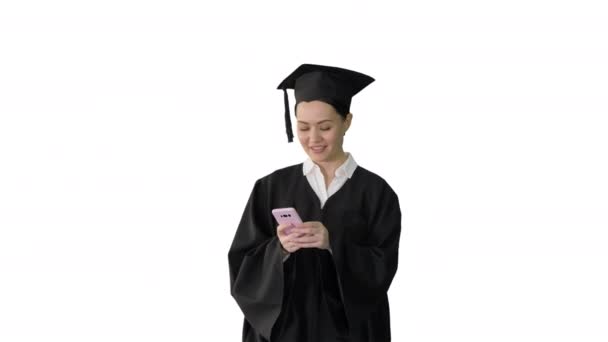 Souriante diplômée en mortier partage de bonnes nouvelles sur son téléphone tout en marchant sur fond blanc. — Video