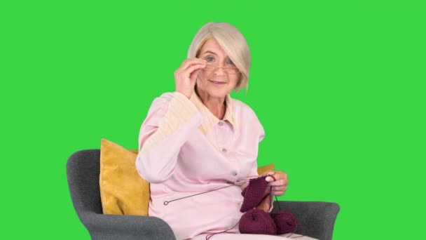 戴眼镜的老妇人在绿色屏幕上编织衣服，并对相机后面的东西做出反应. — 图库视频影像