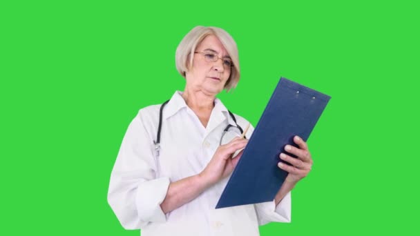 Mature female doctor looking to camera having some serious thoughts and putting down ideas to clipboard on a Green Screen, Chroma Key. — Stock Video