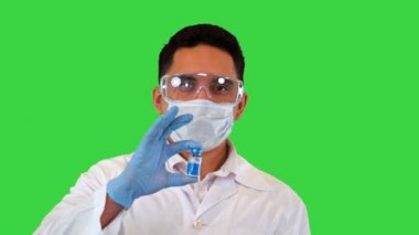 Koruyucu maskeli bir doktor covid-19 aşısı ile bir ampul tutuyor ve yeşil ekran, Chroma Key 'de kameraya gösteriyor..