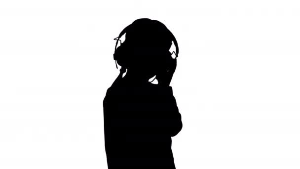 Силуэт Маленькая девочка, одетая в белое, слушает музыку в наушниках и кивает головой в ритм. — стоковое видео