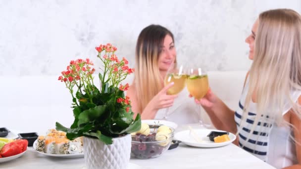 Δύο ελκυστικές κοπέλες, χαρούμενες κολλητές που διασκεδάζουν και πίνουν κρασί στο σπίτι. Δύο ποτήρια λευκό κρασί στα χέρια. Κορίτσια που διασκεδάζουν μαζί κουτσομπολεύοντας. — Αρχείο Βίντεο