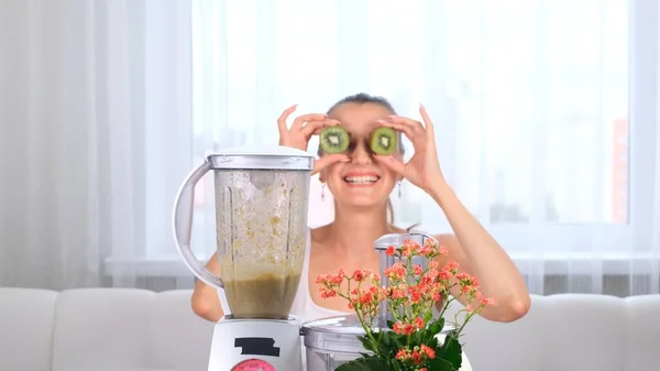 Glückliche junge Frau bereitet köstliche nahrhafte Smoothie im Mixer. Lustiges Weibchen macht Augen aus Kiwi. — Stockfoto
