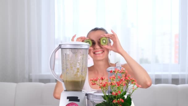 Happy young woman preparing delicious nutritious smoothie in blender. funny female making eyes from kiwi. — Stock Video