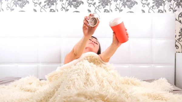 Kobieta wstaje w łóżku z budzikiem i kawą na wynos w rękach, cieszy się porankiem i pije kawę. Koncepcja rozpoczęcia nowego dnia, relaksu w weekend, nie trzeba nigdzie spieszyć — Zdjęcie stockowe