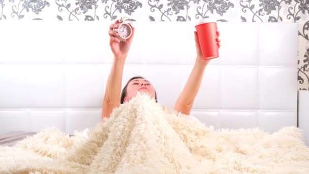 Женщина встает в постели с чашкой кофе и будильником в руках, в ужасе понимает, что она проспала. Концепция нехватки времени, опоздания на работу или в школу — стоковое видео
