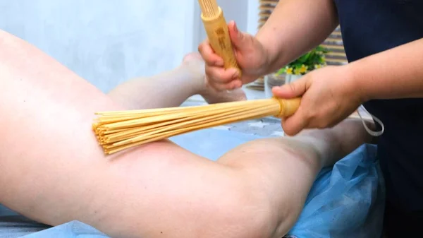 Le masseur fait un massage anti-cellulite des grosses jambes féminines avec des bâtons de bambou. Le thérapeute fait des traitements pour améliorer l'état de la peau. Lutte agressive contre la graisse. Traitement spa. — Photo