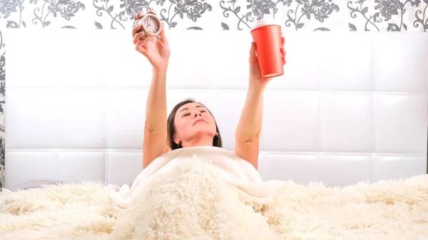 Kobieta wstaje w łóżku z budzikiem i kawą na wynos w rękach, cieszy się porankiem i pije kawę. Koncepcja rozpoczęcia nowego dnia, relaksu w weekend, nie trzeba nigdzie spieszyć — Zdjęcie stockowe
