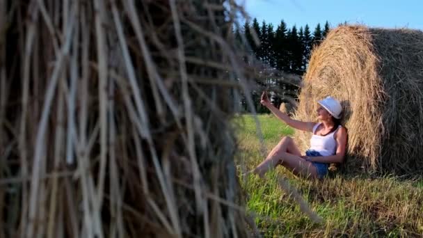 Joven hembra hace una selfie en un teléfono inteligente cerca de un pajar y habla sobre la comunicación de vídeo. Zona rural. concepto de libertad — Vídeo de stock