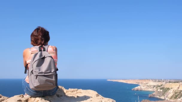 Sırt çantalı mutlu genç bir kadın bir kayanın üzerinde oturur ve aşağıdaki vadiye bakar. Karadeniz 'in kayalık sahillerindeki turistler. Özgürlük ve kolaylık kavramı — Stok video