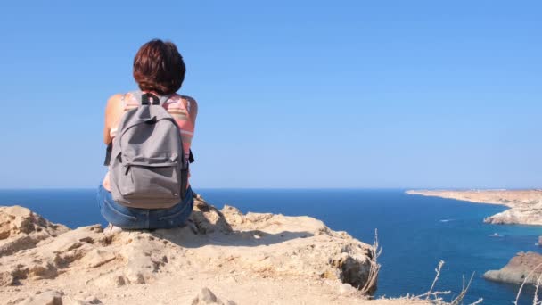 Giovane donna felice con uno zaino si siede su una roccia e guarda la valle sottostante. Turisti sulla costa rocciosa del Mar Nero. Il concetto di libertà e facilità — Video Stock