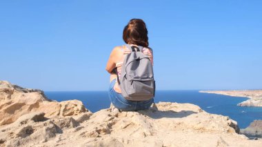 Sırt çantalı mutlu genç bir kadın bir kayanın üzerinde oturuyor ve aşağıdaki vadiye bakıyor. Özgürlük ve rahatlık kavramı.