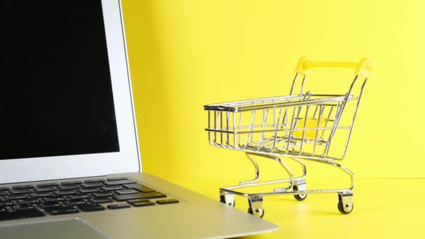 Cesta de compras ao lado de um laptop em um fundo amarelo. Espaço para cópia. Conceito de compras online. — Vídeo de Stock