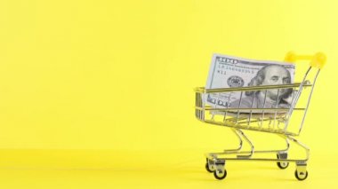 Alışveriş arabası ve sarı arka planda 100 dolarlık banknot, yakın plan. Kara Cuma Alışveriş ve İndirim Konsepti