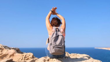 Sırt çantalı genç ve mutlu bir kadın elleri havada bir kayanın üzerinde oturur ve aşağıdaki vadiye bakar. Özgürlük ve kolaylık kavramı.