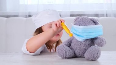 Küçük kız doktorculuk oynuyor, iğne yapıyor. Mutlu çocuk küçük doktor oyuncak ayıyı muayene ediyor. Corona virüsü tedavi konsepti, salgın sırasında enjeksiyon