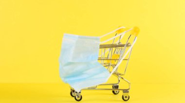 Sarı arka planda alışveriş arabası ve tıbbi koruyucu maske, yakın plan. Kara Cuma Alışveriş ve İndirim Konsepti. Virüslere ve diğer hastalıklara karşı korunma kavramı, tıp.