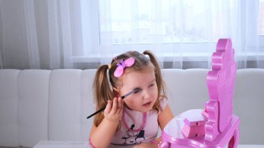 Pembe elbiseli ve boncuklu sevimli küçük kız aynanın önünde makyajla oynuyor..