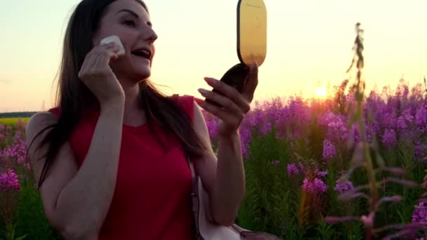 Jonge vrouwelijke brunette tussen de wilg thee bloeiwijzen bij zonsondergang poeders haar gezicht, zet op make-up en kijkt in de spiegel. Meisje in hoge stelen van vuurwier op een heldere zonnige avond — Stockvideo