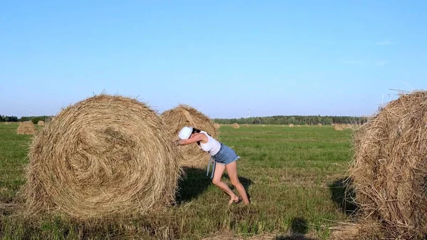 Beautiful young girl in a hat pushes a haystack. Rural area. Feminist strong woman concept — Stock Photo, Image