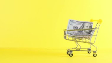 Alışveriş arabası ve sarı arka planda 100 dolarlık banknot, yakın plan. Kara Cuma Alışveriş ve İndirim Konsepti