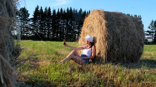 Young female makes a selfie on a smartphone near a haystack and talks on video communication. Rural area. freedom concept — Stock Photo, Image