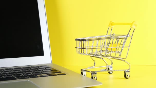 Bevásárlókosár mellett egy laptop sárga háttér. Másold a helyet. Online vásárlási koncepció. — Stock videók