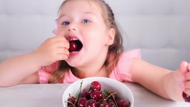 Bella bambina carina seduta a un tavolo di legno leggero con un piatto di ciliegie e mangiare una bacca. Note di alimentazione. — Video Stock