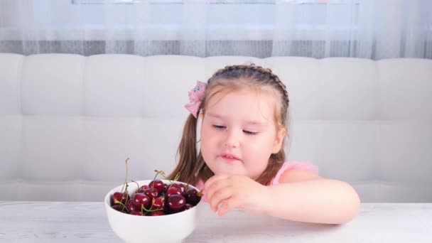 Vacker söt blond liten flicka sitter vid ett ljust träbord med en tallrik körsbär och äter röda bär. Hälsosam kost. — Stockvideo