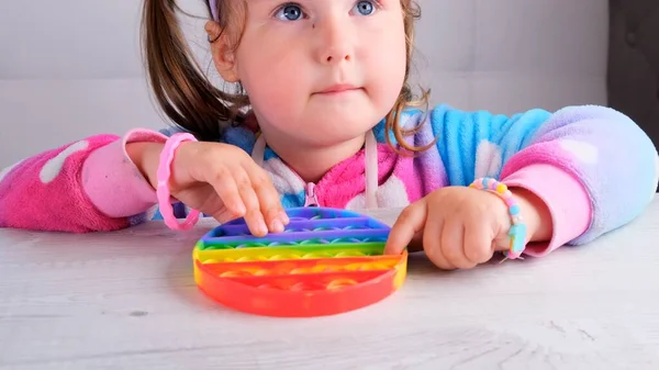 可爱的小女孩带着弹出它的感觉玩具圆圈的形式。小雌蜂在白色的背景上挤压着色彩斑斓的彩虹状的柔软硅胶气泡。缓解压力和焦虑。时尚的坐立不安游戏. — 图库照片