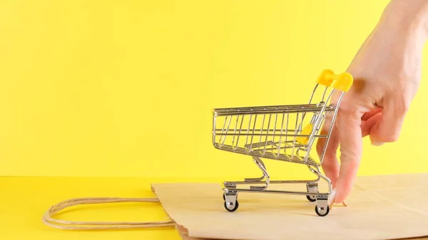 Mano femenina empuja un juguete carrito de compras sobre un fondo amarillo, primer plano. Viernes Negro Compras y descuento Concepto — Foto de Stock