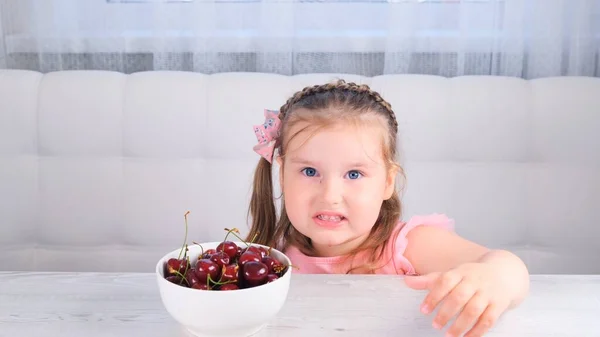 Beautiful smiling little girl eating cherries over white. little cute girl sitting with a plate of cherries and eating a berry. — Stock Photo, Image