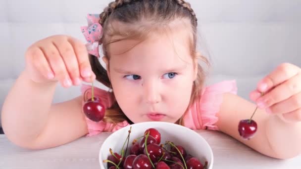 Söt liten flicka sitter vid ett ljust träbord med en tallrik körsbär och håller röda bär. Hälsosam kost. — Stockvideo