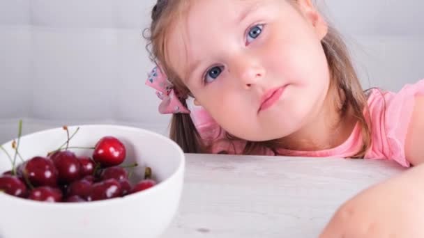 Portret van een schattig klein meisje zittend aan een lichte houten tafel met een bord kersen en het eten van een bes. Gezond eten. — Stockvideo