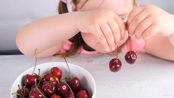 Cute little girl sitting at a light wooden table with a plate of cherries and holding red berries. Healthy eating. — Stock Photo, Image