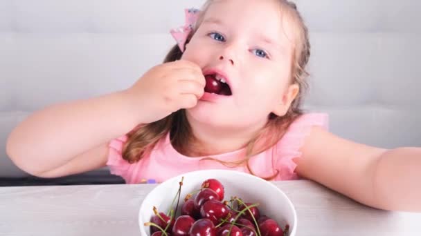 Mooi blond klein meisje zittend aan een lichte houten tafel met een bord kersen en het eten van rode bessen. Gezond eten. — Stockvideo