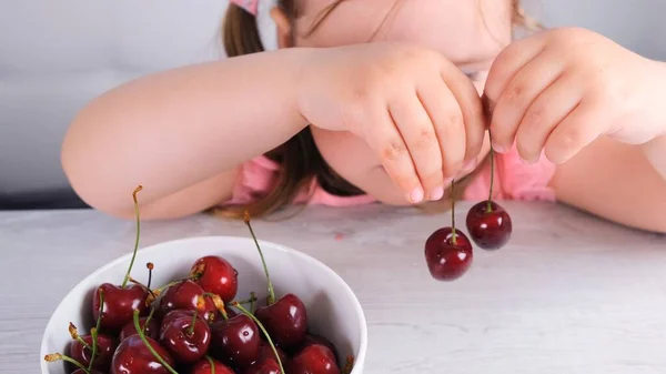 Pretty little girl sitting at a light wooden table with a plate of cherries and holding red berries. Healthy eating. — Stock Photo, Image