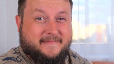 Şık saç stili ve sakallı mutlu genç adam kameraya bakıyor, alaycı bir şekilde gülümsüyor ve şaşırıyor..