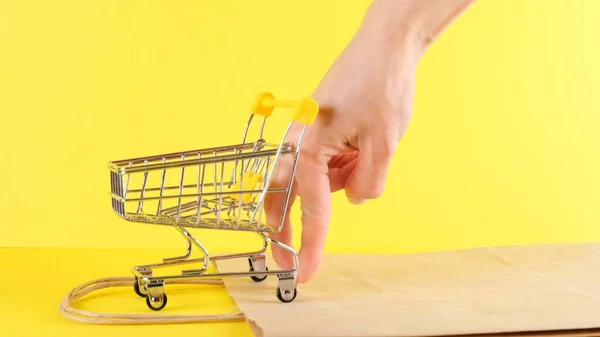 Kvinnlig hand skjuter en leksak Shopping vagn på en gul bakgrund, närbild. Black Friday Shopping och rabatterat koncept — Stockfoto