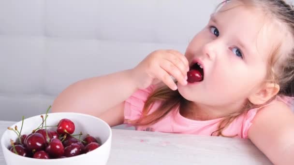 Vacker söt liten flicka sitter vid ett ljust träbord med en tallrik körsbär och äter ett bär. Hälsosam kost. — Stockvideo