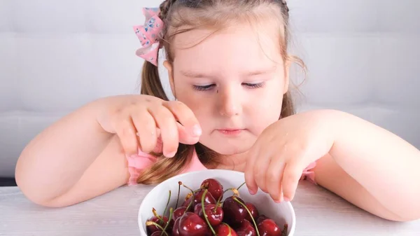 Beautiful cute blonde little girl sitting at a light wooden table with a plate of cherries and eating red berries. Healthy eating. — Stock Photo, Image