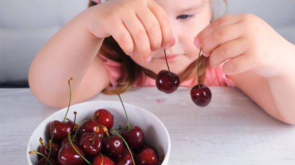 Aranyos kislány ül egy könnyű fa asztalnál egy tányér cseresznyével és kezében vörös bogyók. Egészséges táplálkozás. — Stock Fotó