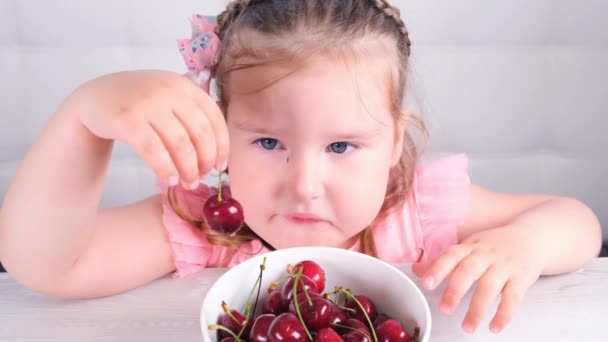 Mooi klein meisje zitten aan een lichte houten tafel met een bord kersen en met rode bessen. Gezond eten. — Stockvideo