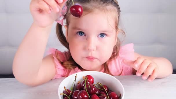 Söt blond liten flicka sitter vid ett ljust träbord med en tallrik körsbär och äter röda bär. Hälsosam kost. — Stockvideo
