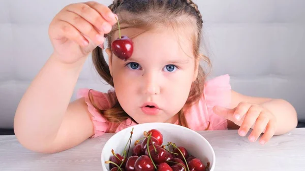 Bonita niña rubia sentada en una mesa de madera clara con un plato de cerezas y la celebración de bayas rojas. Alimentación saludable. — Foto de Stock
