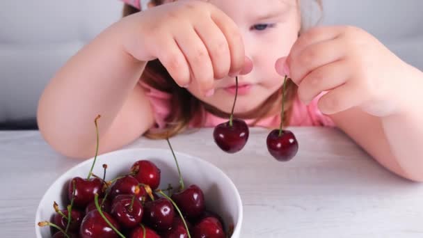 Bonita niña rubia sentada en una mesa de madera clara con un plato de cerezas y la celebración de bayas rojas. Alimentación saludable. — Vídeos de Stock