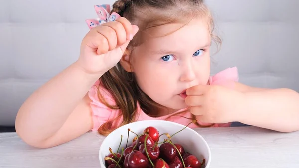 Cute blonde little girl sitting at a light wooden table with a plate of cherries and eating red berries. Healthy eating. — Stock Photo, Image