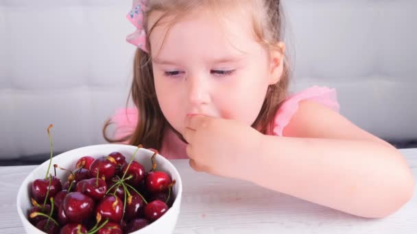 Bella sorridente bambina mangiare ciliegie sopra bianco. piccola ragazza carina seduta con un piatto di ciliegie e mangiare una bacca. — Video Stock