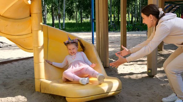 Vrolijke moeder die haar dochter beneden van de glijbaan ziet glijden. Grappige familieactiviteit in het park, plezier hebben. Happy childhood concept: Moskou, Rusland - 18 augustus 2021 — Stockfoto