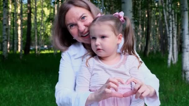 Happy loving grandmother and granddaughter, 3 years old, show their heart out of their hands. The concept of a happy relationship between generations, raising children — Stock Video